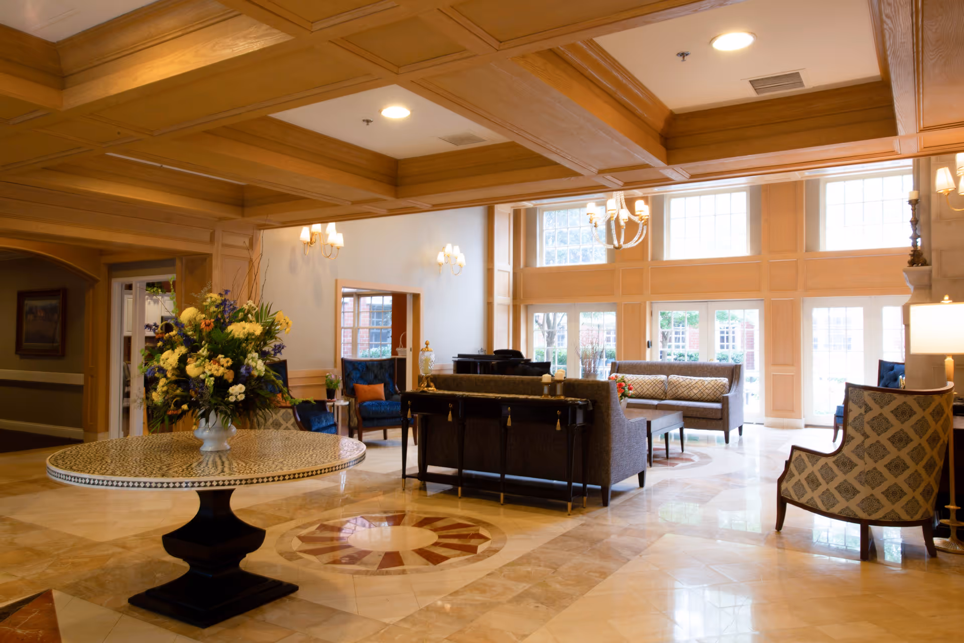 A spacious and elegant living room area with high ceilings and large windows allowing natural light to fill the space. The room features a round table with a large floral arrangement in the foreground, several upholstered chairs and sofas arranged around a coffee table, and warm wood paneling on the ceiling and walls. There are chandeliers and wall sconces providing additional lighting.