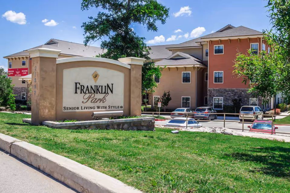 Exterior view of a senior living facility with a large sign in the foreground that reads 'Franklin Park Senior Living With Style'. The building is multi-story with beige and reddish-brown walls, stone accents, and several windows. There are cars parked in the parking lot and a tree providing some shade. The sky is blue with a few clouds.