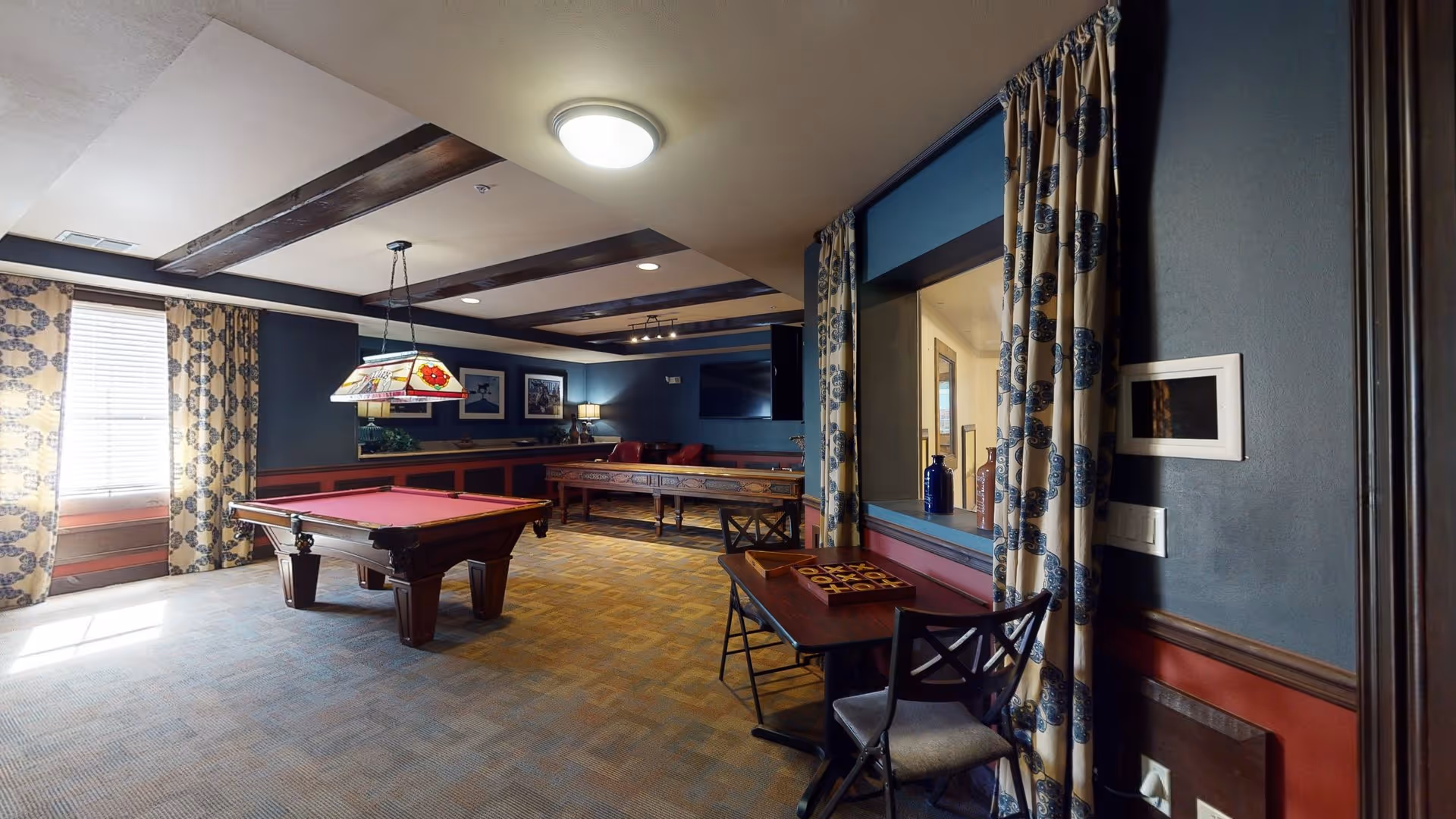 A recreational room with a pool table in the center, a shuffleboard table against the far wall, and a small table with chairs and a tic-tac-toe game near a window with patterned curtains. The room has blue walls with red wainscoting, exposed wooden ceiling beams, and framed pictures on the walls. There is a ceiling light fixture and a hanging lamp above the pool table.