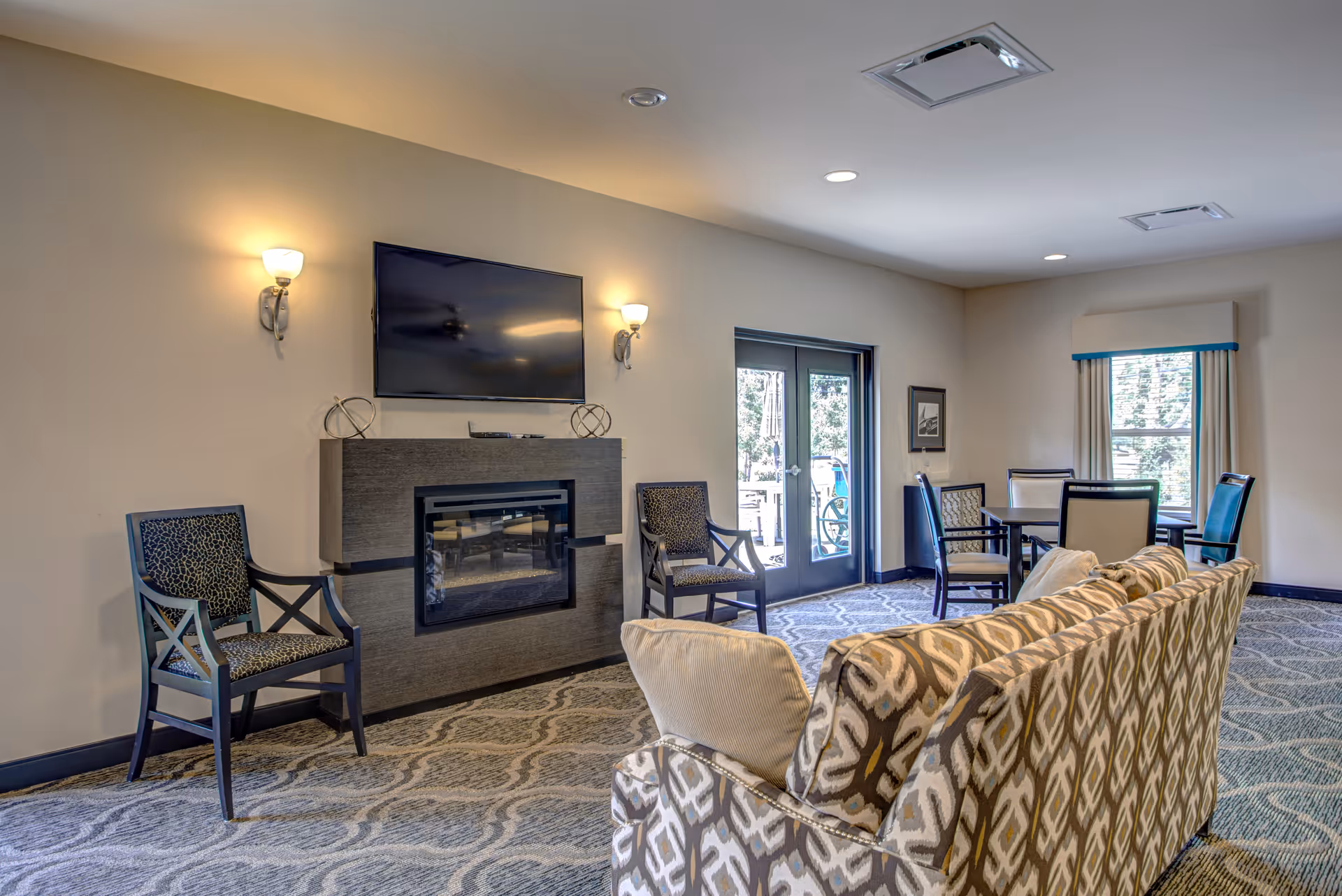 A cozy living room area in a senior living facility featuring a patterned sofa with cushions, two armchairs with patterned upholstery, a modern fireplace with a flat-screen TV mounted above it, and a small dining table with four chairs near a window and glass doors leading outside. The room is well-lit with wall sconces and recessed ceiling lights.