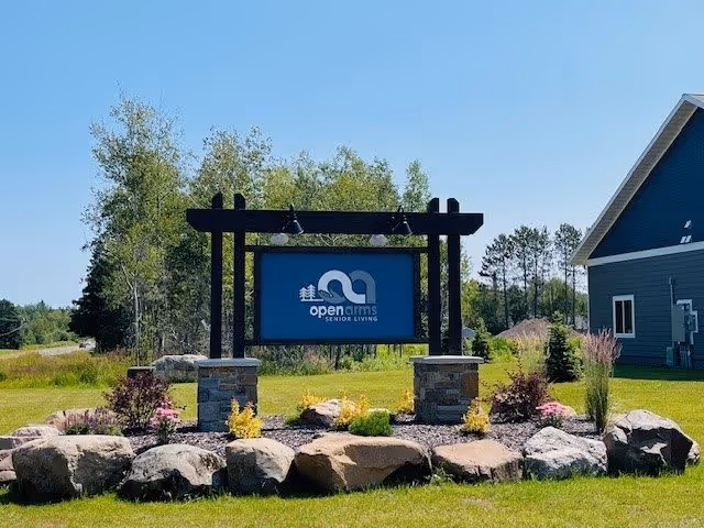 Outdoor view of a sign for Open Arms Senior Living mounted on a black wooden frame with stone pillars at the base, surrounded by rocks and landscaped plants, with a blue sky and trees in the background and a blue building partially visible on the right.
