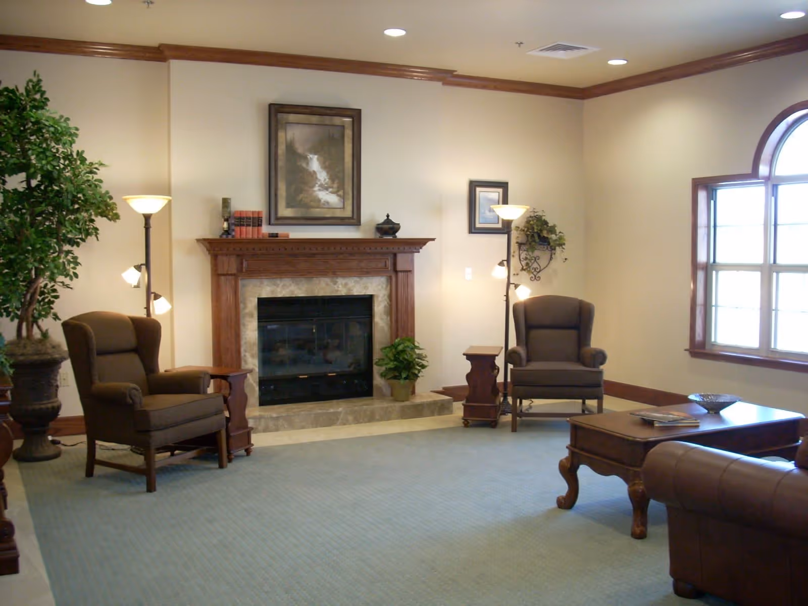 A cozy living room with two brown armchairs, a wooden coffee table, a fireplace with a wooden mantel, framed artwork above the fireplace, two floor lamps, a large potted plant, and a window with an arched top letting in natural light.
