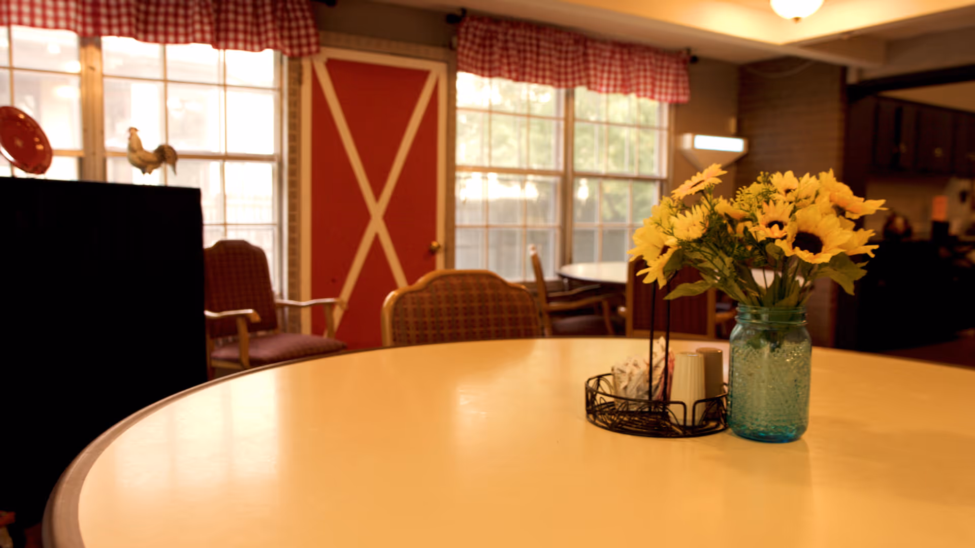 Round dining table with a vase of sunflowers and seating in a cozy common area with large windows and a red door.