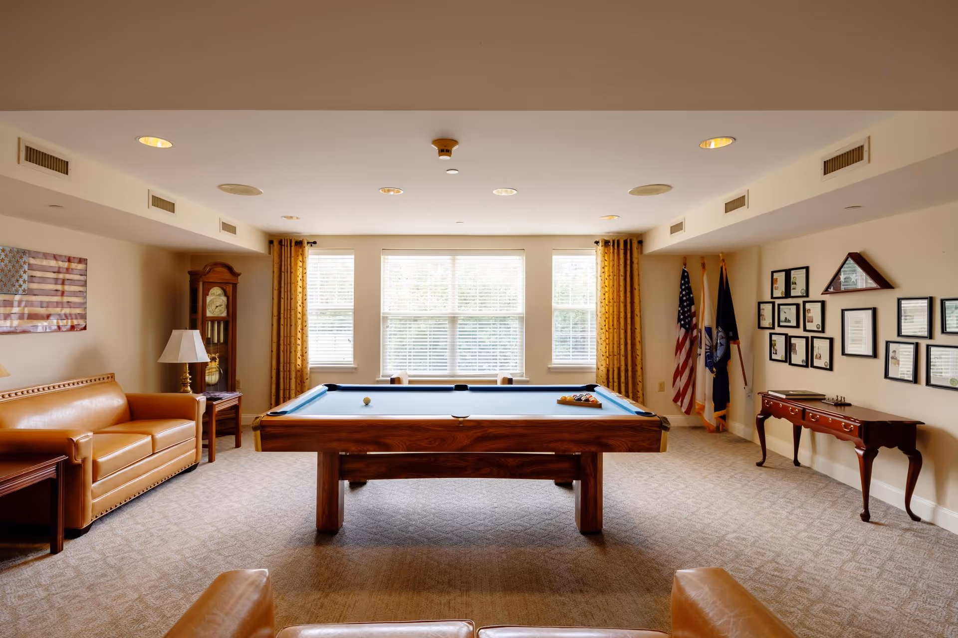 A well-lit recreational room with a pool table in the center, surrounded by leather sofas and chairs. The room has large windows with yellow curtains, an American flag and other flags in the corner, a grandfather clock, framed certificates and memorabilia on the wall, and a wooden side table with a lamp.