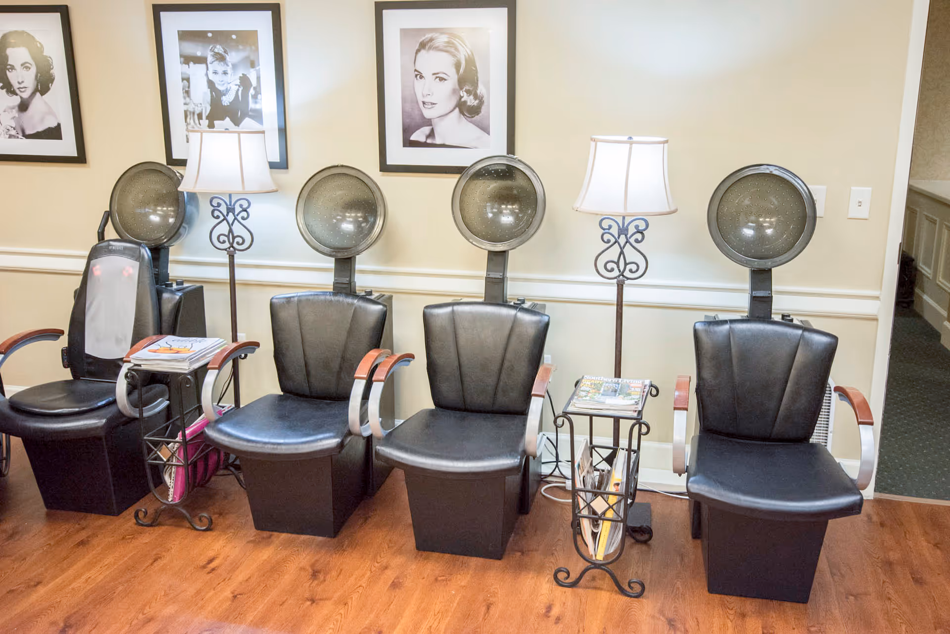 A row of four black salon chairs with hair dryers above them in a room with wooden flooring. Two floor lamps with white shades are placed between the chairs. On the wall behind the chairs are three framed black and white portraits of women. There are small side tables with magazines next to the chairs.