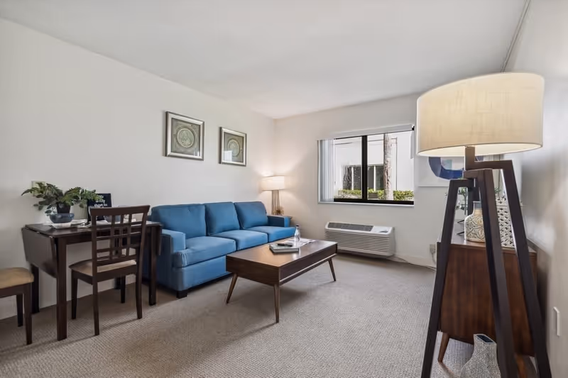 A cozy living room with a blue sofa, a wooden coffee table with a water bottle and book on it, a small dining table with two chairs, a floor lamp, and a window with a view of the outside. The room has light-colored walls and carpeted floor.