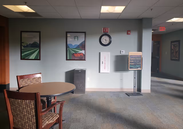 A quiet indoor common area with a round table and two chairs with patterned upholstery. The wall behind has two framed landscape posters, a clock, a fire extinguisher cabinet, and a small signboard with meal times and a positive message. The floor is carpeted and there is an exit door visible down a hallway to the right.