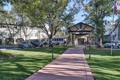 A paved walkway leading to the entrance of a senior living facility building named Winslow Court Assisted & Senior Living, surrounded by green lawns, trees, parked cars, and an American flag on a flagpole.