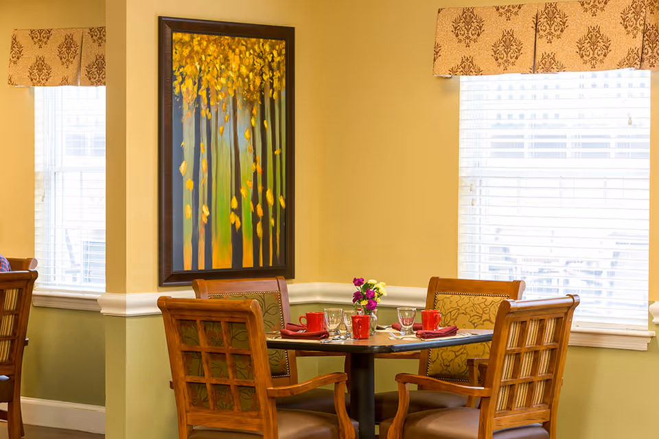 A dining area with a round table set for four people, featuring red cups, glasses, and napkins. The room has two windows with white blinds and patterned valances. A framed painting of trees with yellow leaves hangs on the wall between the windows. Four wooden chairs with patterned cushions surround the table.