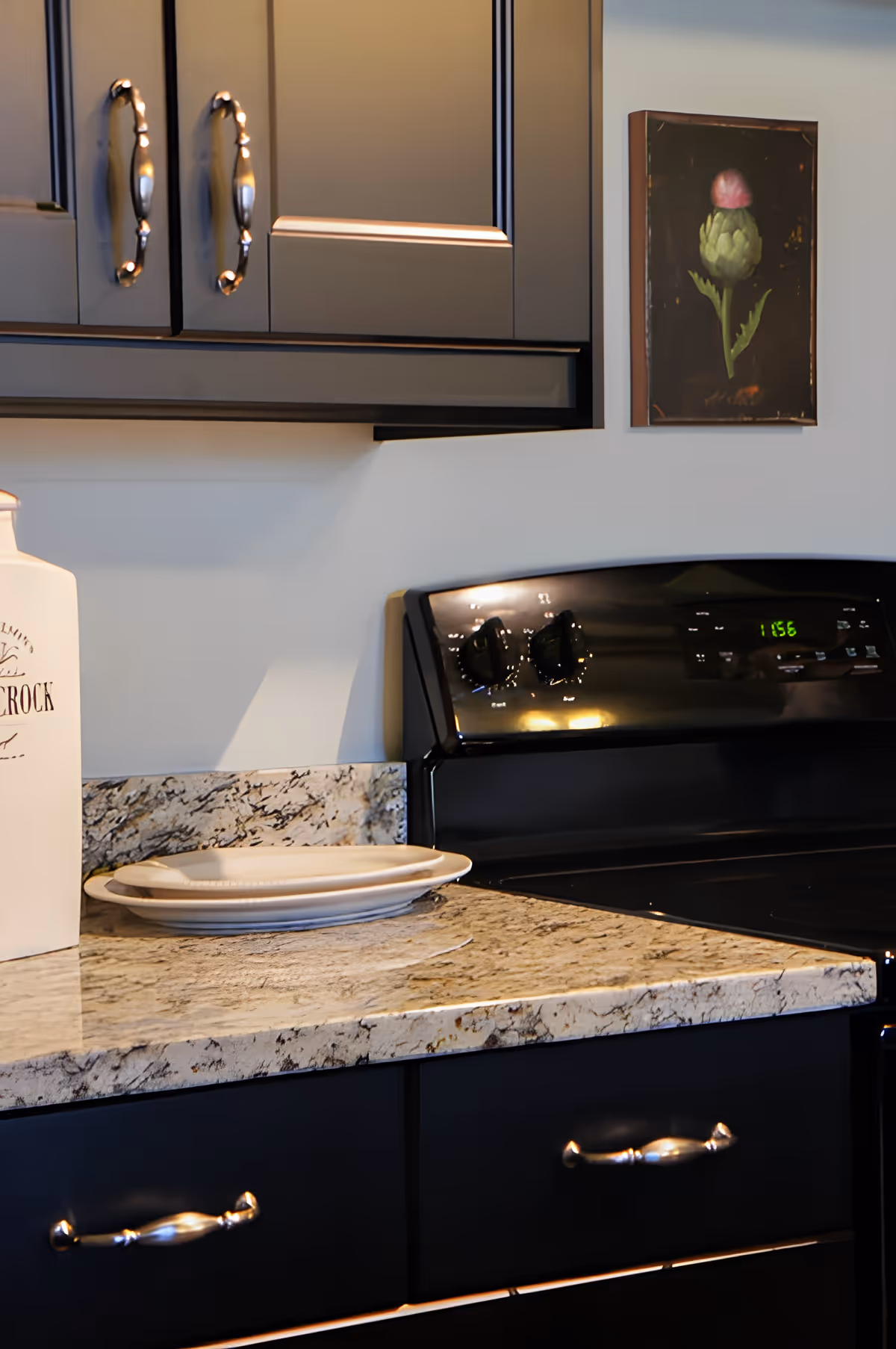 Granite kitchen countertop with a black stove, dark cabinets, stacked plates and a small framed floral painting on the wall.