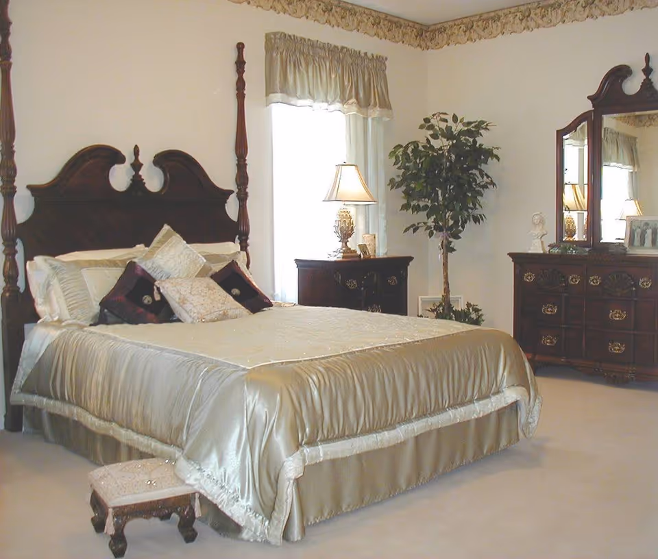 A bedroom with a large wooden four-poster bed covered in a shiny beige bedspread and multiple pillows. There is a small upholstered footstool at the foot of the bed. To the right, there is a wooden dresser with a large mirror and decorative items on top. A nightstand with a lamp and a potted plant are positioned near a window with beige curtains.