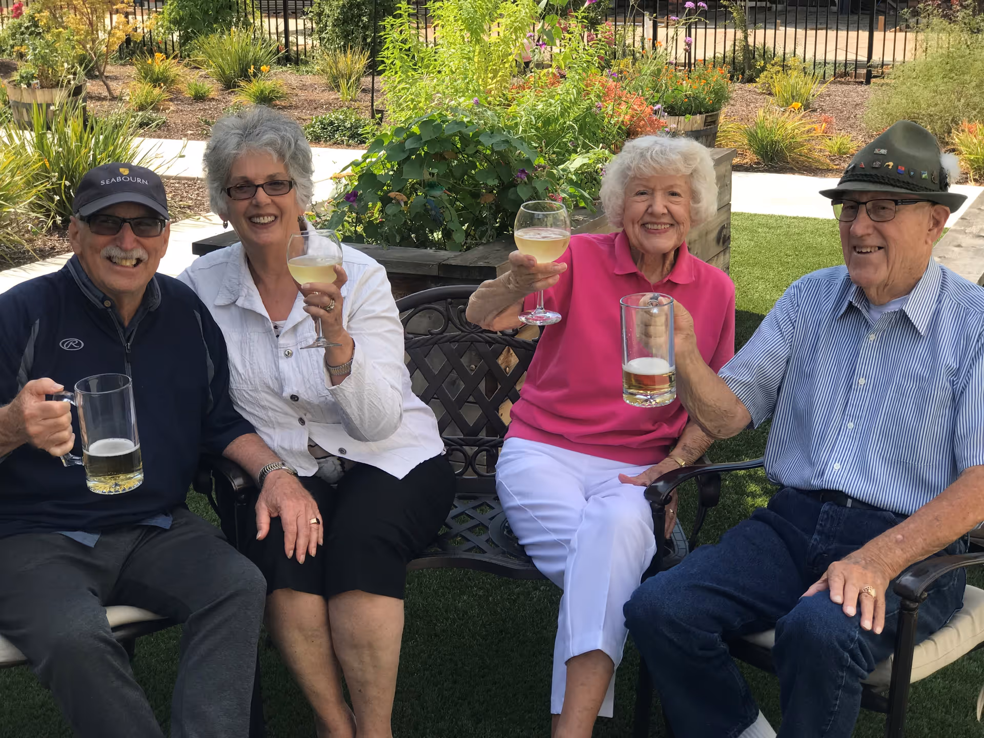 Four elderly individuals sitting outdoors on chairs and a bench, smiling and holding up glasses of beer and wine in a celebratory toast. They are surrounded by greenery and garden plants in a sunny setting.