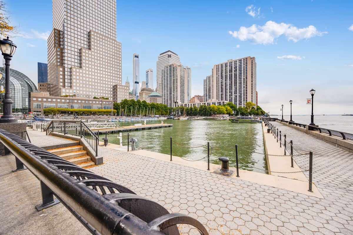 A waterfront promenade with a paved walkway, black metal railings, and street lamps. There are steps leading down to a dock area with moored boats. In the background, there are tall buildings and trees under a partly cloudy sky.