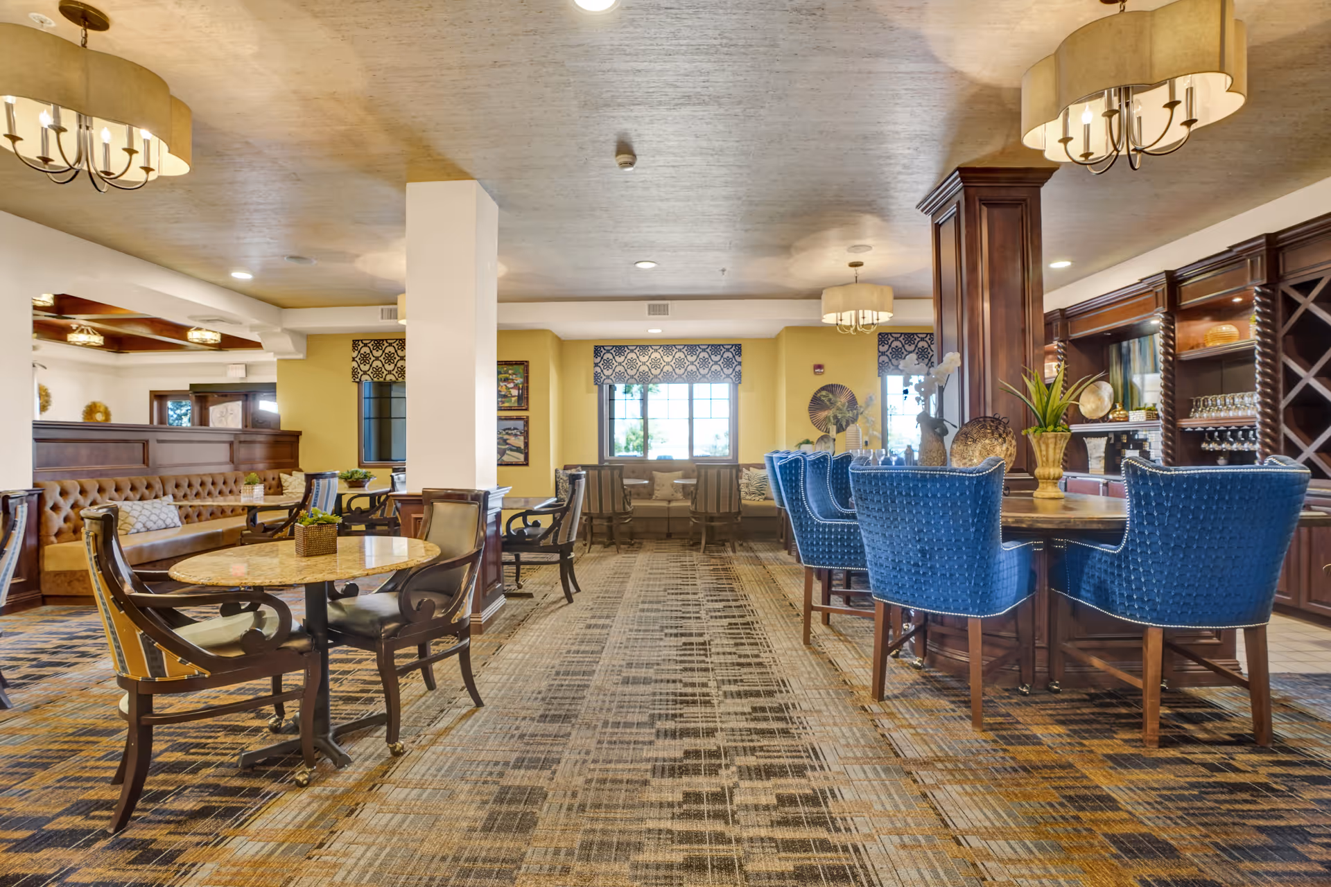 A spacious and well-lit common area in a senior living facility featuring multiple seating arrangements including a round table with chairs, a long cushioned bench along the wall, and a bar area with blue upholstered chairs. The room has large windows with patterned valances, warm yellow walls, and decorative lighting fixtures hanging from the ceiling. There are plants and decorative items on the tables and shelves, creating a welcoming atmosphere.