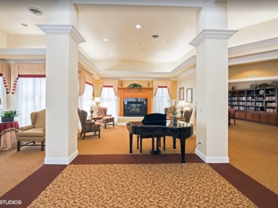 A spacious and well-lit living room area with a grand piano in the center, surrounded by comfortable armchairs and a fireplace with a wooden mantel. Large windows with curtains allow natural light to fill the room, and a bookshelf filled with books is visible in the background.