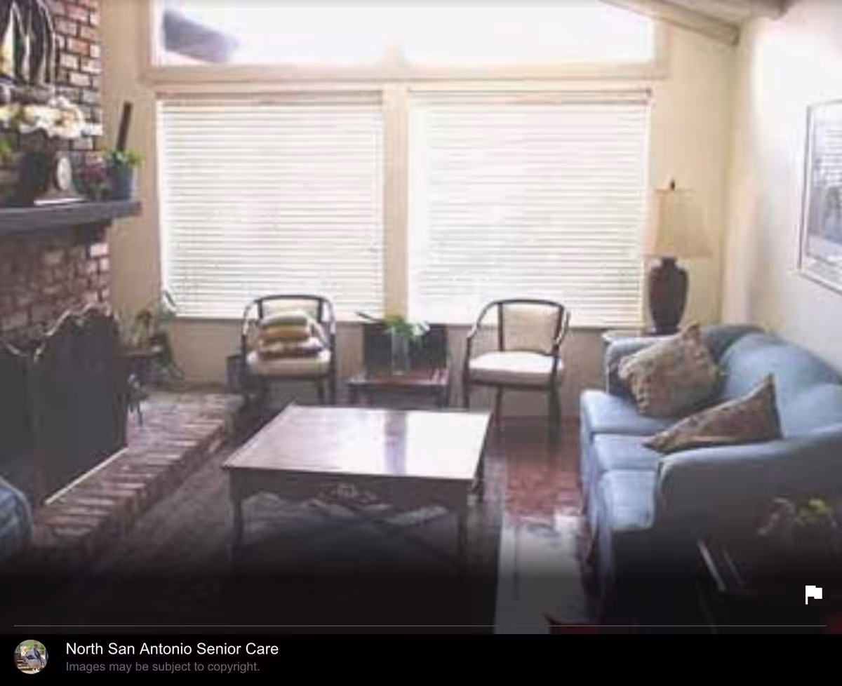 A cozy living room with a blue sofa adorned with pillows, two chairs with cushions, a wooden coffee table, a brick fireplace with a mantel decorated with plants and a clock, and large windows with blinds allowing natural light to fill the room.