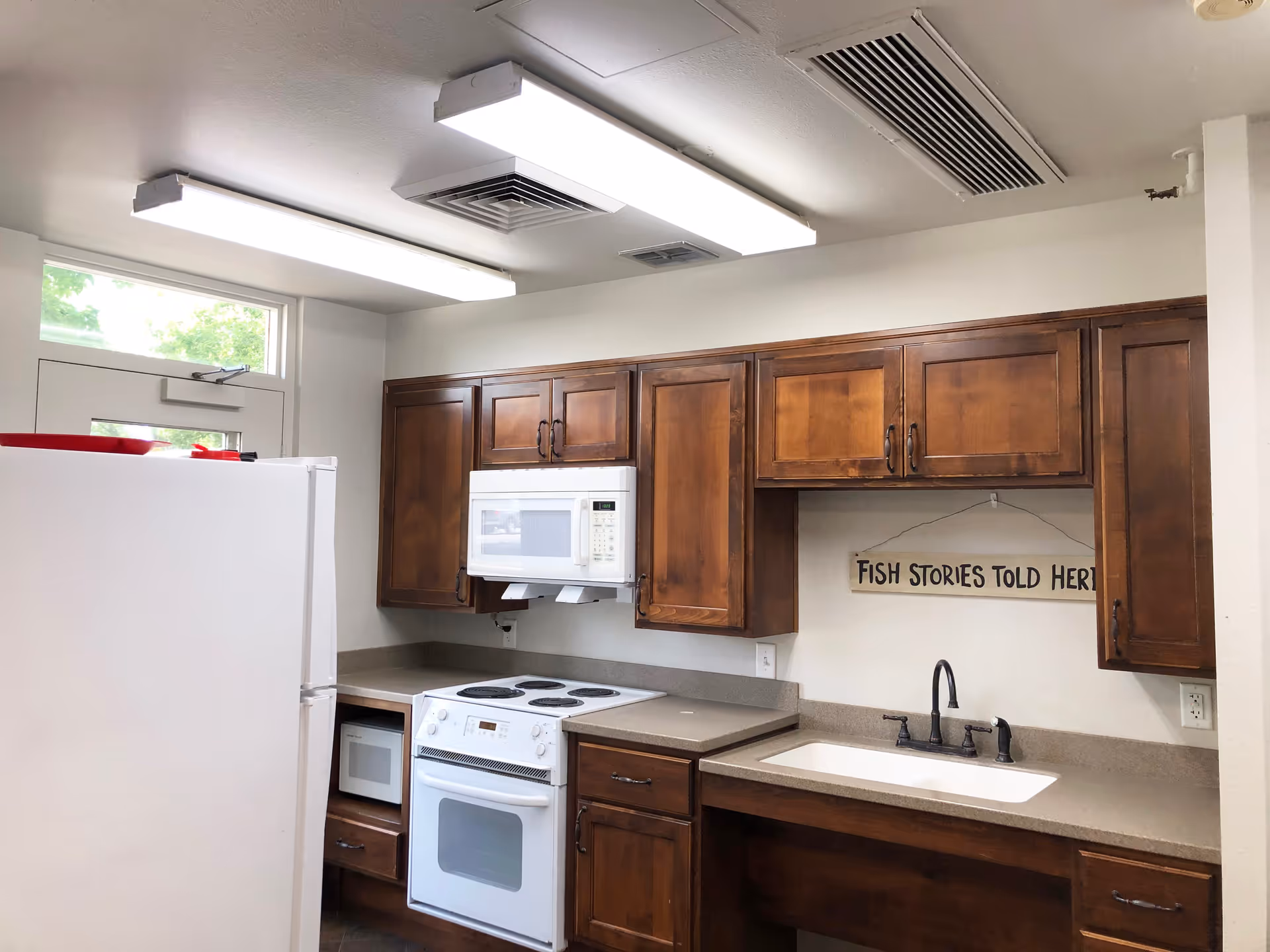 Interior view of a kitchen with wooden cabinets, a white refrigerator, a white microwave above a white stove, a sink with a black faucet, and a sign on the wall that reads 'FISH STORIES TOLD HERE'. There is a window above the door letting in natural light and fluorescent ceiling lights.