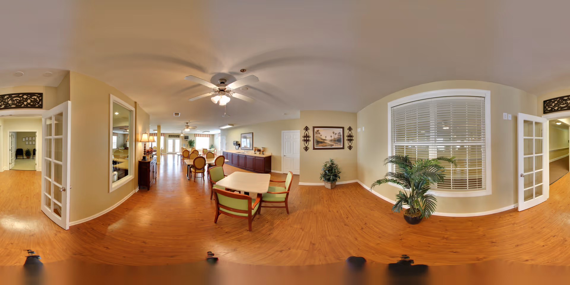 A spacious, well-lit common area in a senior living facility with wooden flooring, beige walls, and ceiling fans. The room features multiple tables and chairs arranged for dining or socializing, decorative plants, framed artwork on the walls, and large windows with blinds. French doors lead to adjacent rooms and hallways.