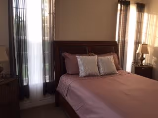 A bedroom with a wooden bed frame and a neatly made bed with a mauve bedspread and three decorative pillows. There are two windows with sheer curtains allowing natural light to enter the room. A bedside table with a lamp is visible on the right side of the bed.