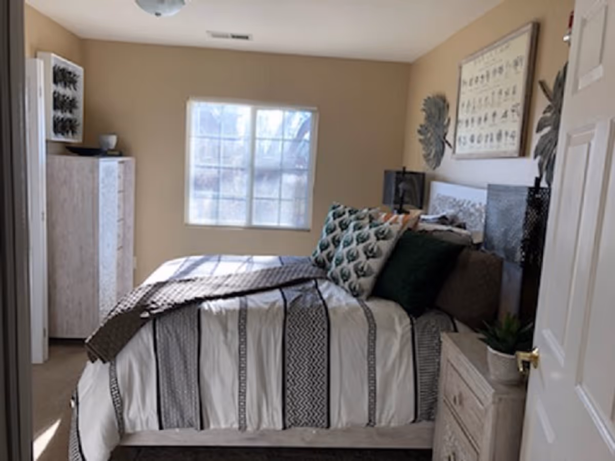 A cozy bedroom with a bed covered in a white and black striped comforter and multiple decorative pillows. There is a window letting in natural light, a dresser on the left side, and a nightstand with a small plant on the right. The walls are beige with framed artwork and decorative metal wall hangings.