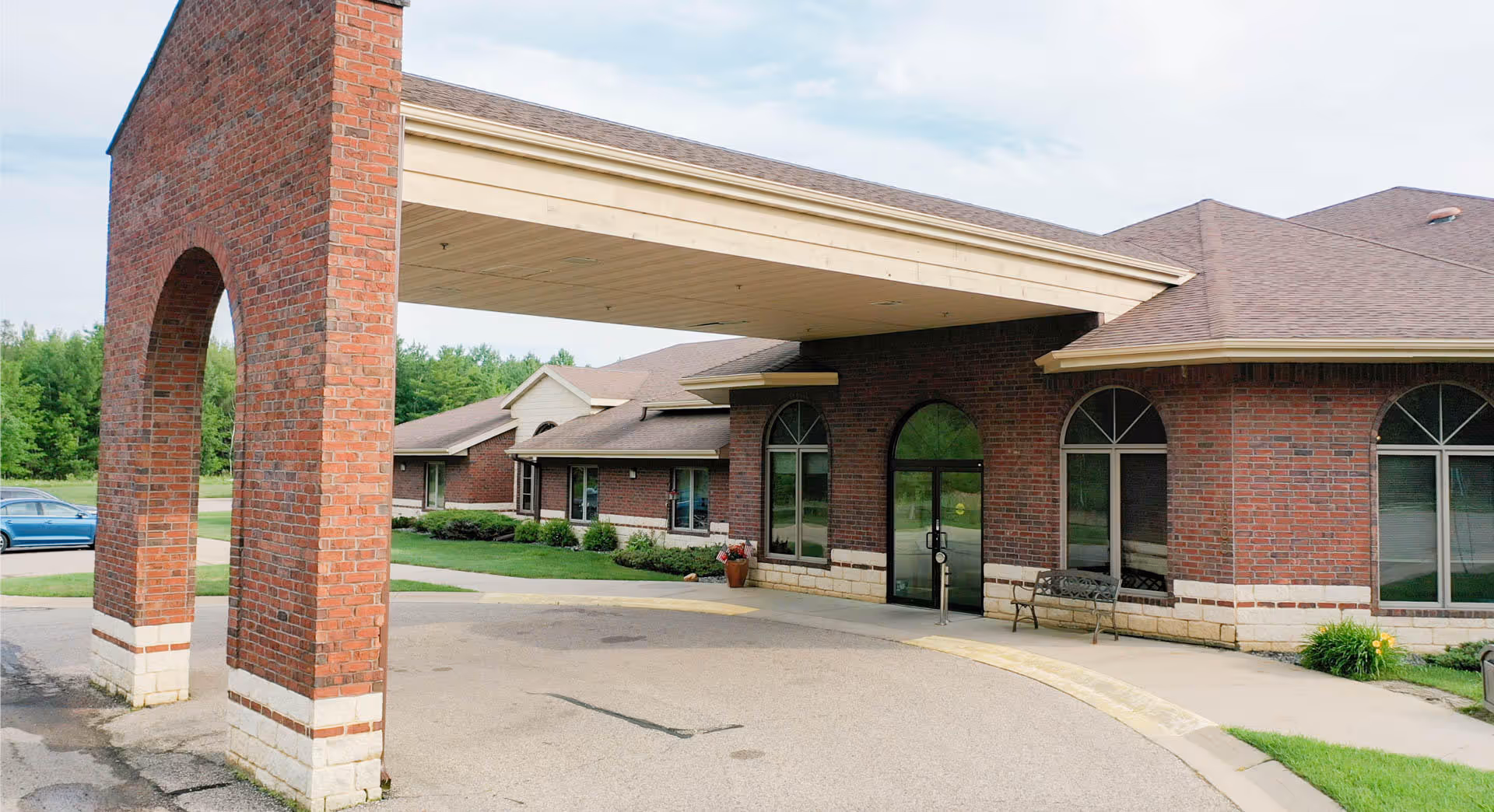 Covered drive-up entrance of a brick senior living building with arched windows and a bench outside the glass doors.