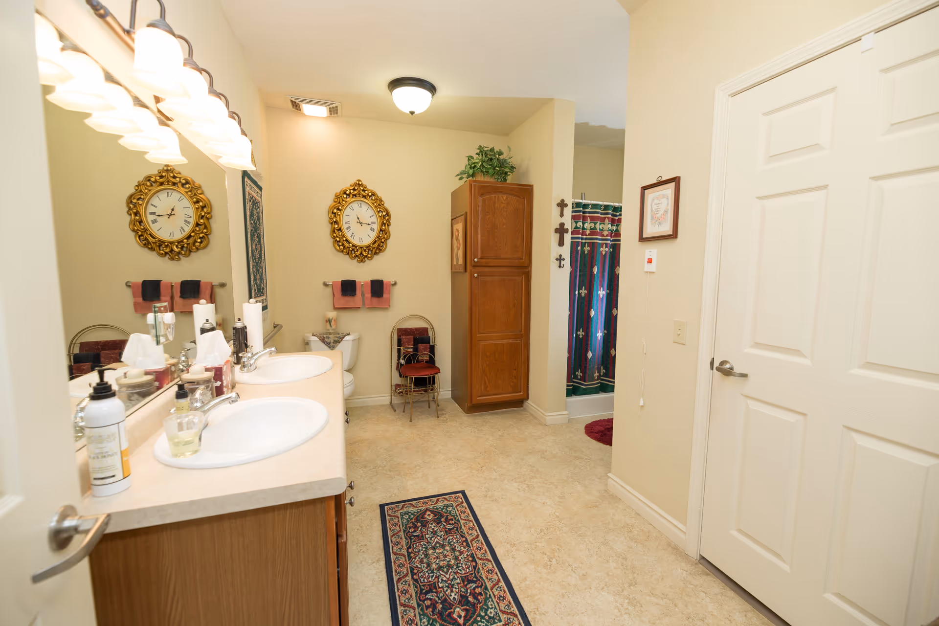 A well-lit bathroom with a double sink vanity on the left, a large mirror above the sinks, and a decorative wall clock. There is a toilet with towels hanging above it, a wooden cabinet with a plant on top, and a shower with a colorful curtain in the background. The floor has a patterned rug, and the walls are painted beige with some framed decorations.