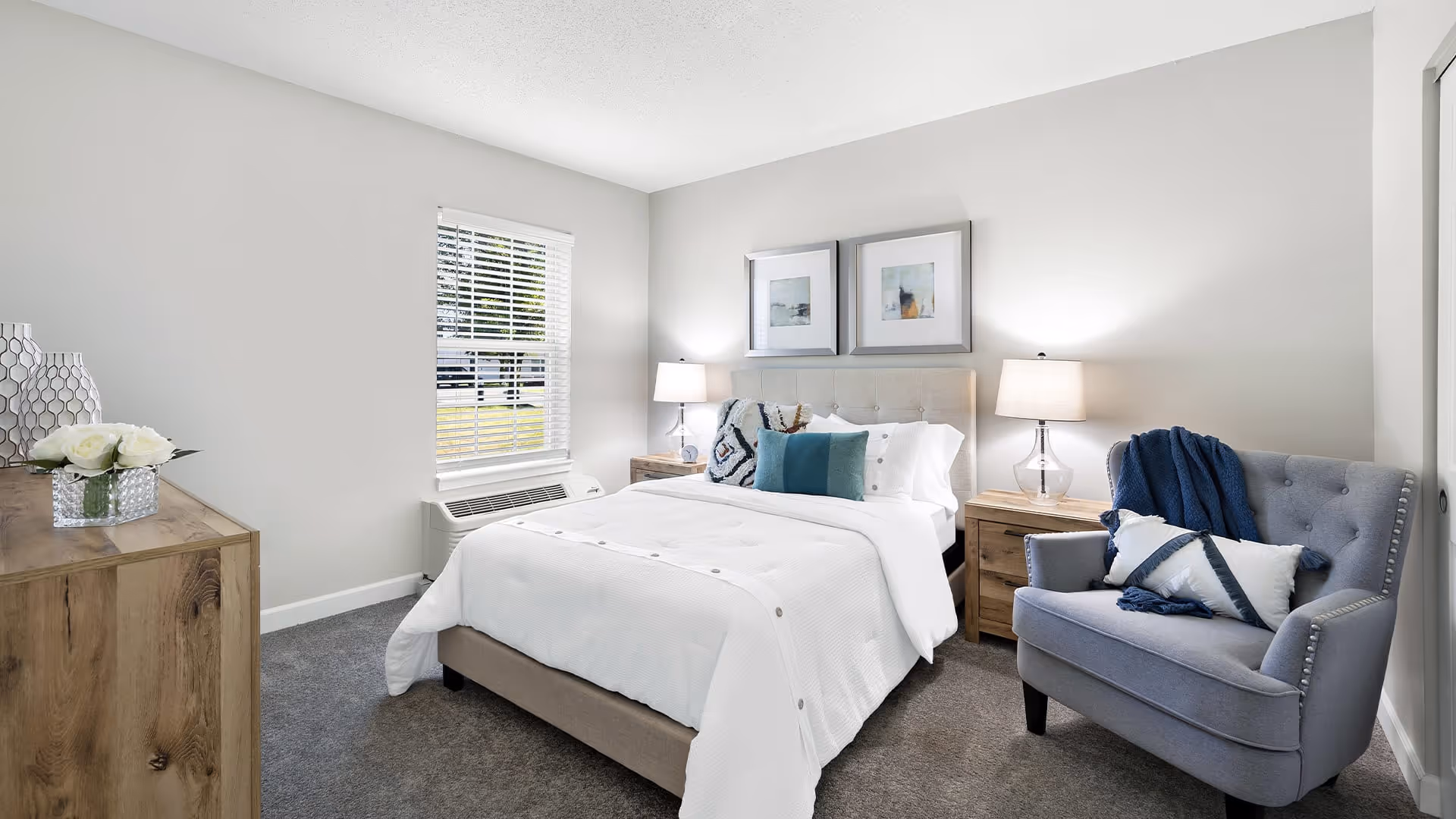 A neatly made bed with white bedding and decorative pillows in a bright bedroom. The room features two wooden nightstands with lamps, a gray upholstered armchair with a blue throw blanket and pillow, a wooden dresser with decorative items, a window with white blinds, and two framed pictures on the wall above the bed.
