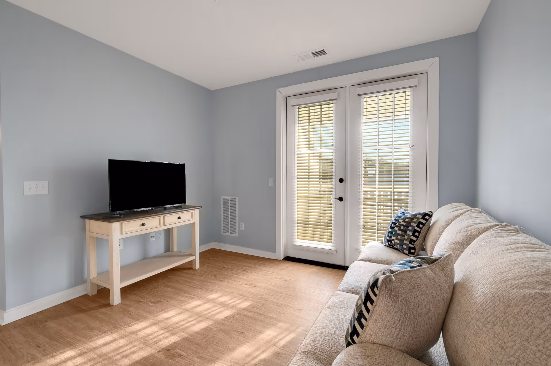 A small living room with light blue walls and wooden flooring. There is a beige couch with patterned cushions on the right side and a white wooden TV stand with a flat-screen TV on the left. French doors with blinds lead outside, allowing natural light to enter the room.