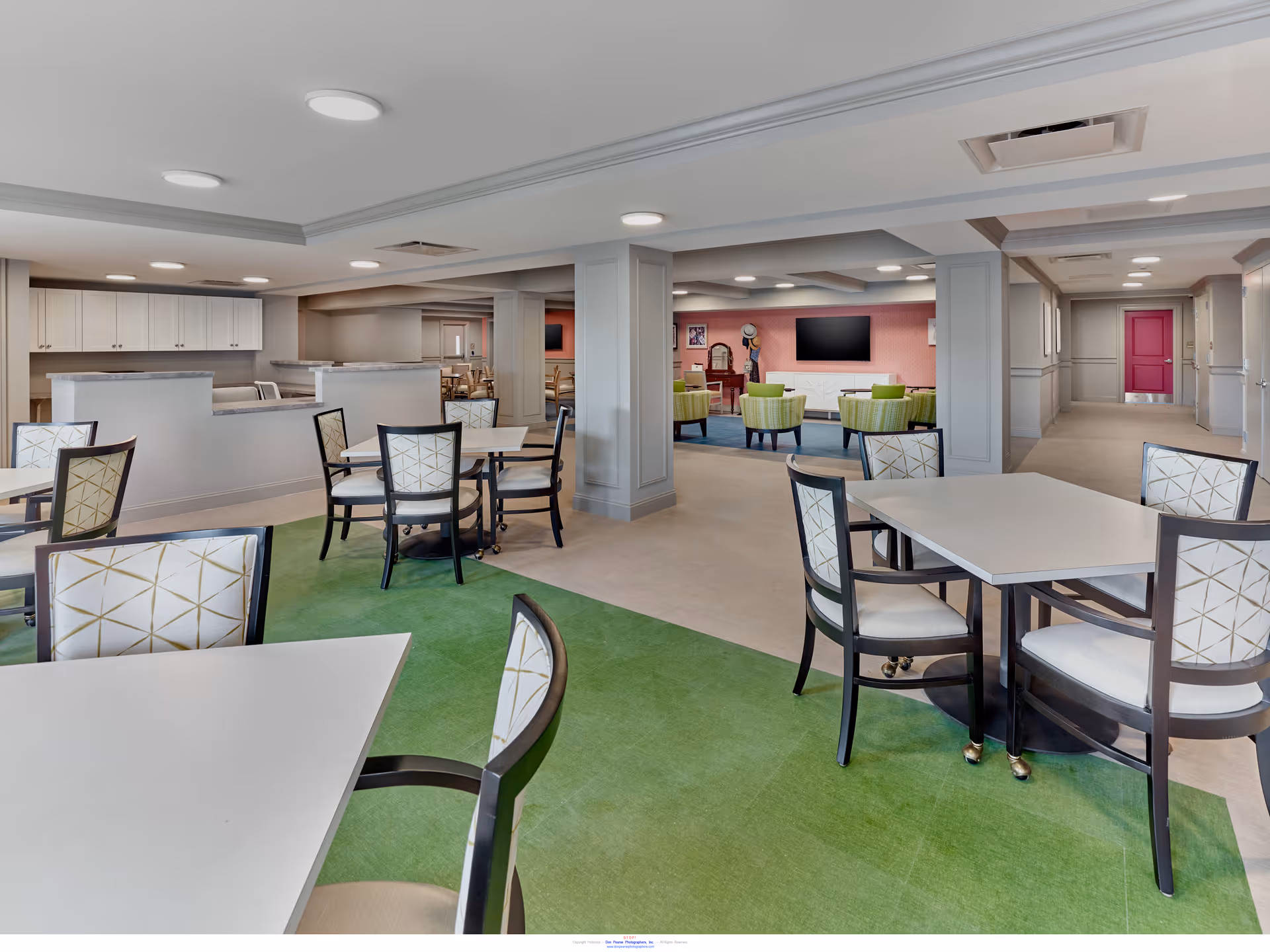 A spacious and well-lit common area in a senior living facility featuring multiple tables and chairs with patterned upholstery on a green carpet. In the background, there is a lounge area with green armchairs, a large flat-screen TV mounted on a pink wall, and a hallway with a red door at the end.