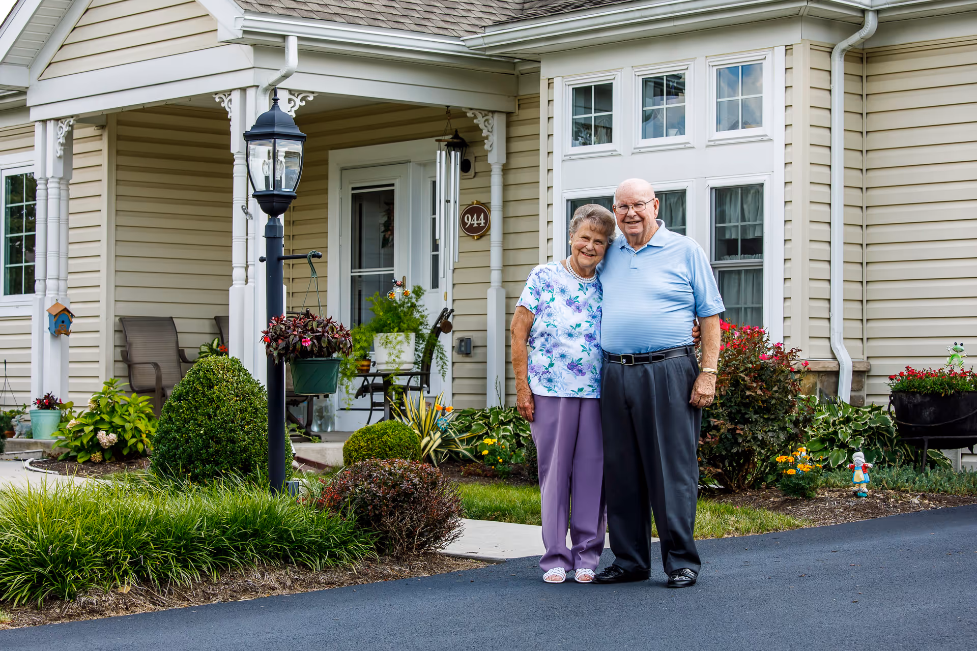 An elderly couple standing arm in arm outside a beige residential building with a porch, surrounded by well-maintained garden plants and flowers.