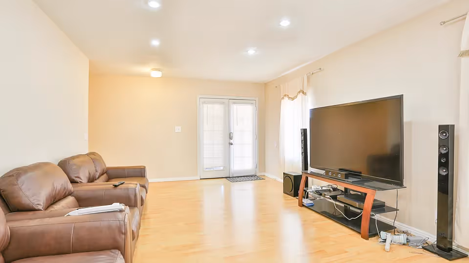 A bright living room with light wooden flooring, beige walls, and recessed ceiling lights. The room features two brown leather recliners on the left side and a large flat-screen TV on a black and wooden TV stand on the right side, flanked by two tall black speakers. French doors with blinds and a window with curtains allow natural light into the space.