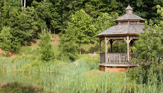 A wooden gazebo with a shingled roof situated near a pond surrounded by tall grass and trees in a lush green outdoor setting.