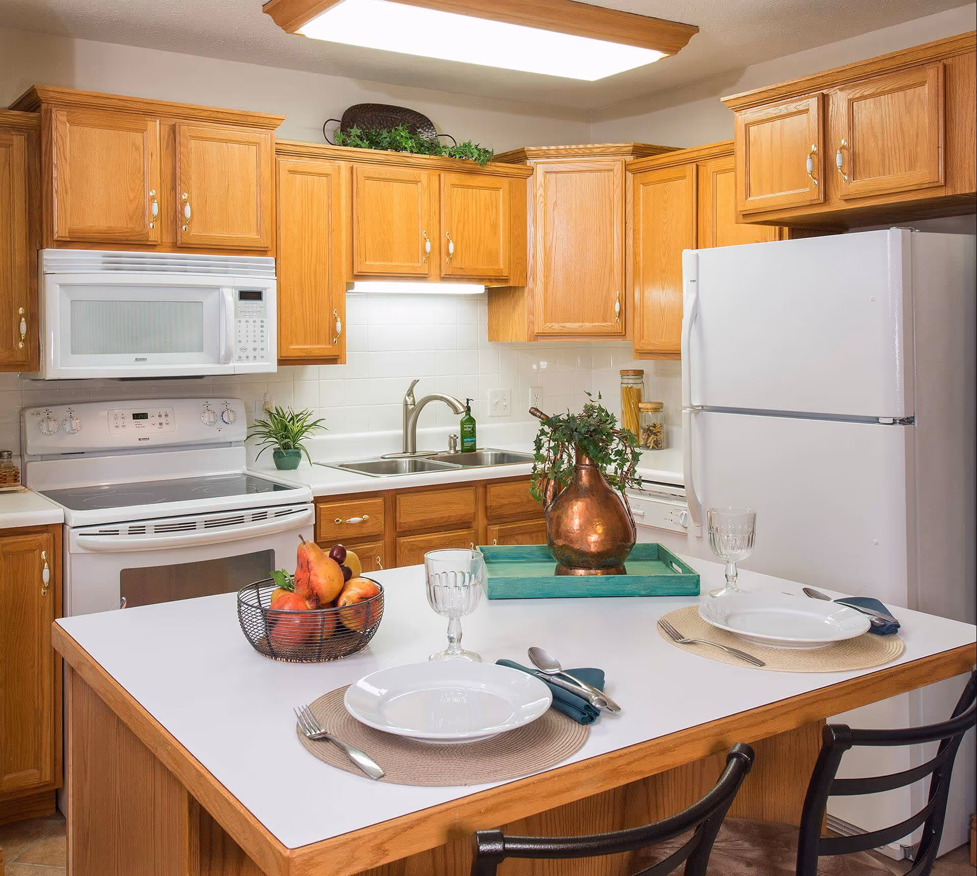 Bright kitchen with oak cabinets, white appliances, and an island set for two with a fruit bowl and plant centerpiece.