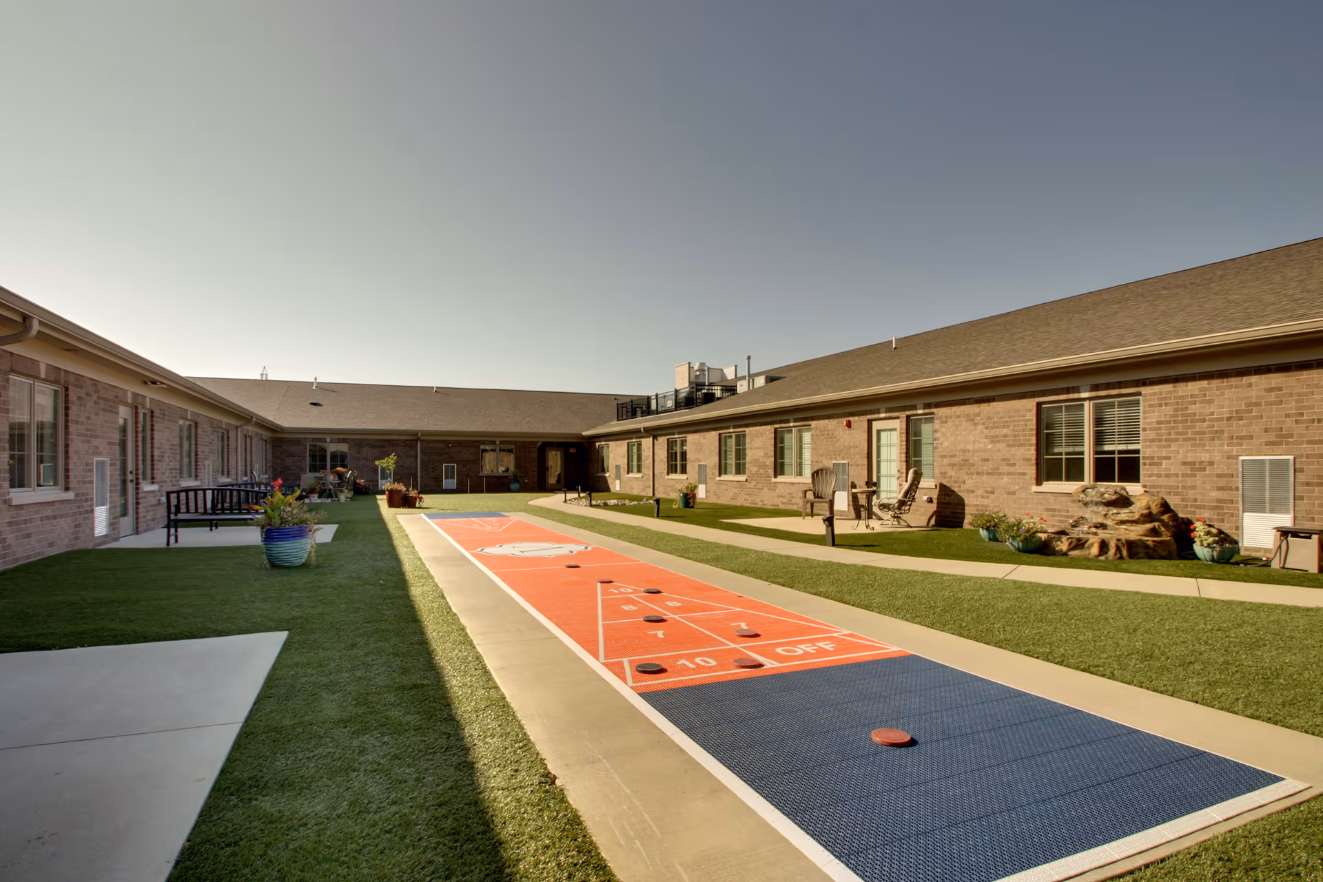 Outdoor courtyard area at Evergreen Senior Living featuring a shuffleboard court with several shuffleboard pucks on it. The courtyard is surrounded by single-story brick buildings with windows and doors opening onto the courtyard. There are benches, potted plants, and chairs placed along the walkways beside the shuffleboard court under a clear sky.