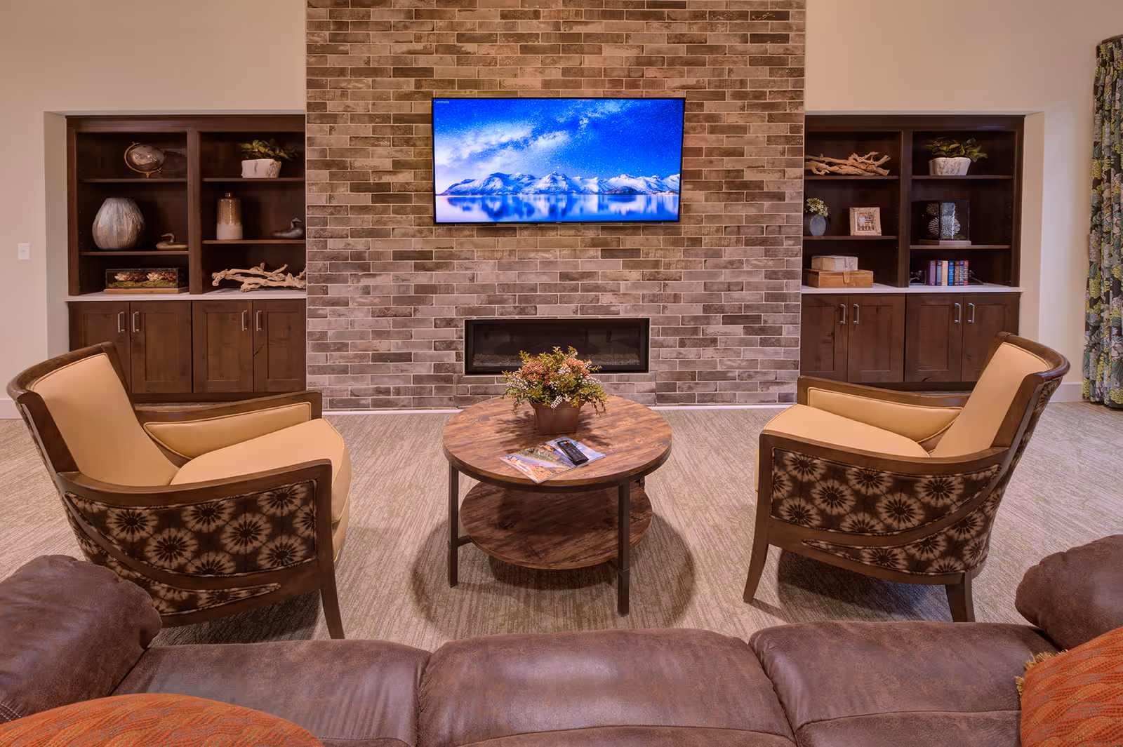 Cozy communal living room with two armchairs facing a wall-mounted TV above a brick fireplace and a round coffee table.