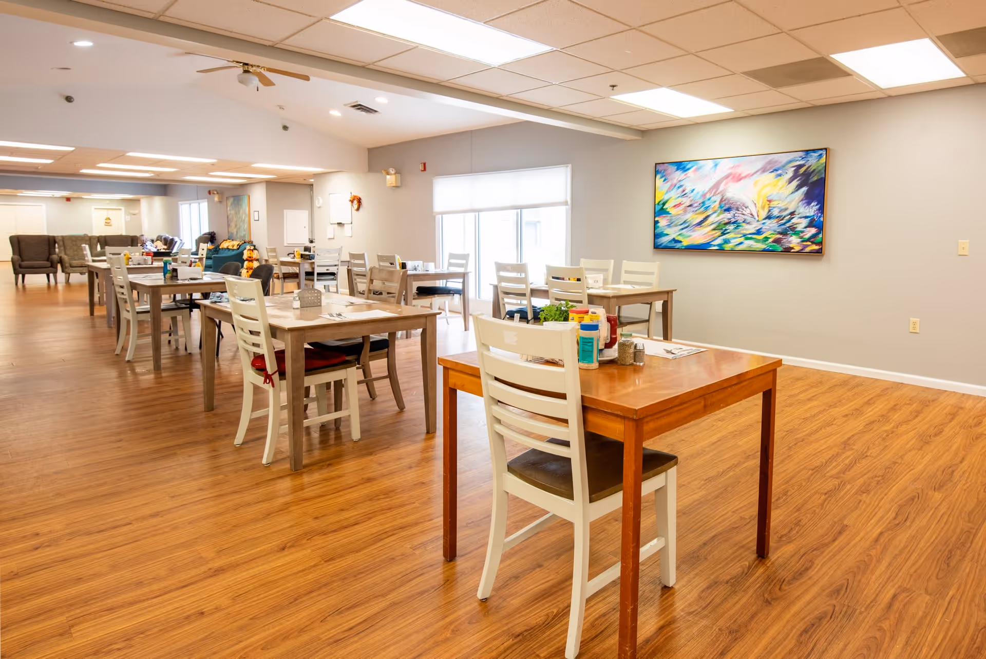 A spacious dining room with multiple wooden tables and white chairs arranged neatly. The room has wooden flooring, a ceiling fan, and a large colorful abstract painting on the wall. There are windows allowing natural light to brighten the space, and some tables have condiments and napkins on them.