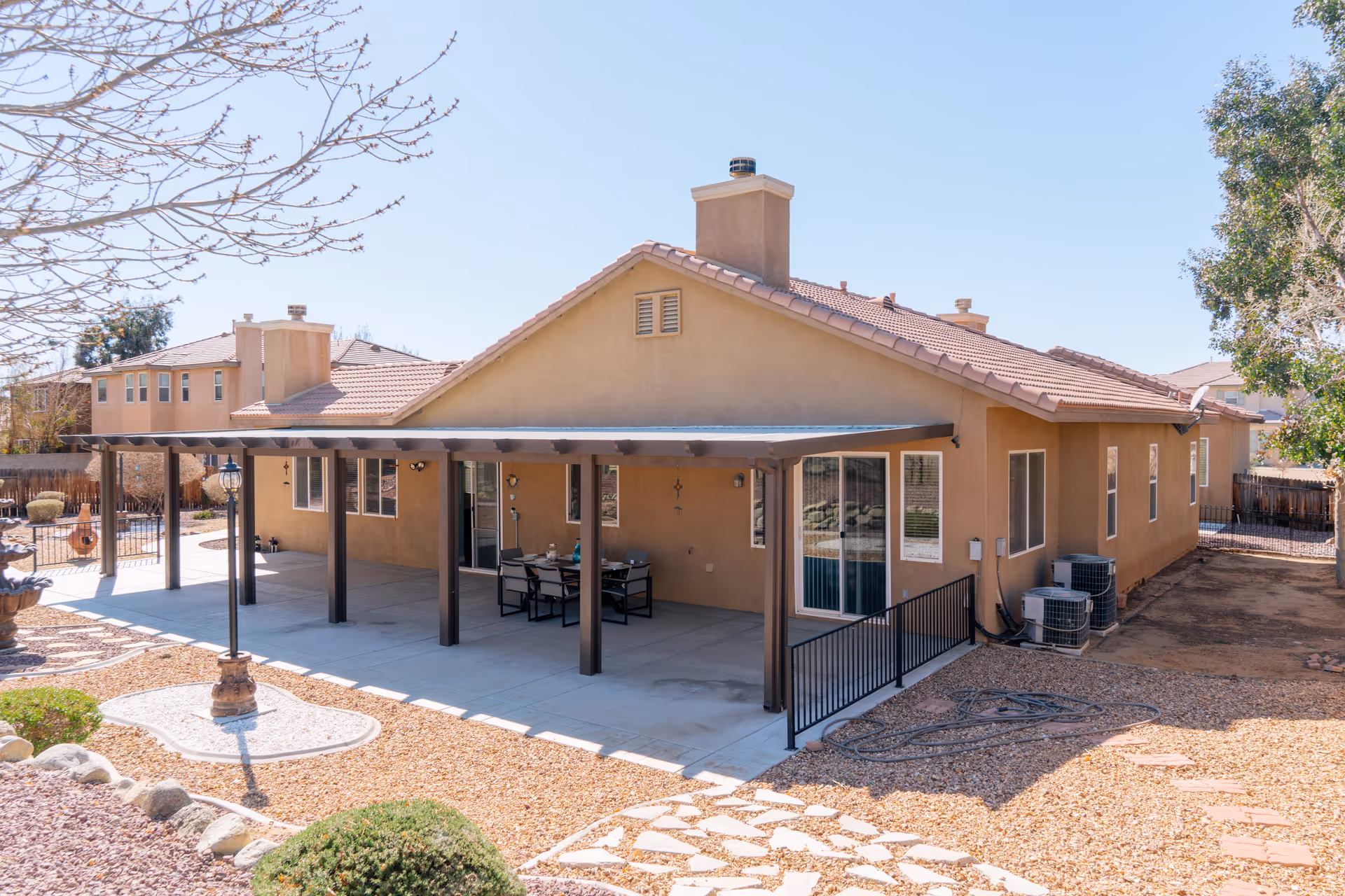 Back exterior view of a single-story beige stucco house with a covered patio area supported by dark wooden posts. The patio has a table and chairs, and the yard features gravel landscaping with some bushes and a stone pathway. The sky is clear and blue.