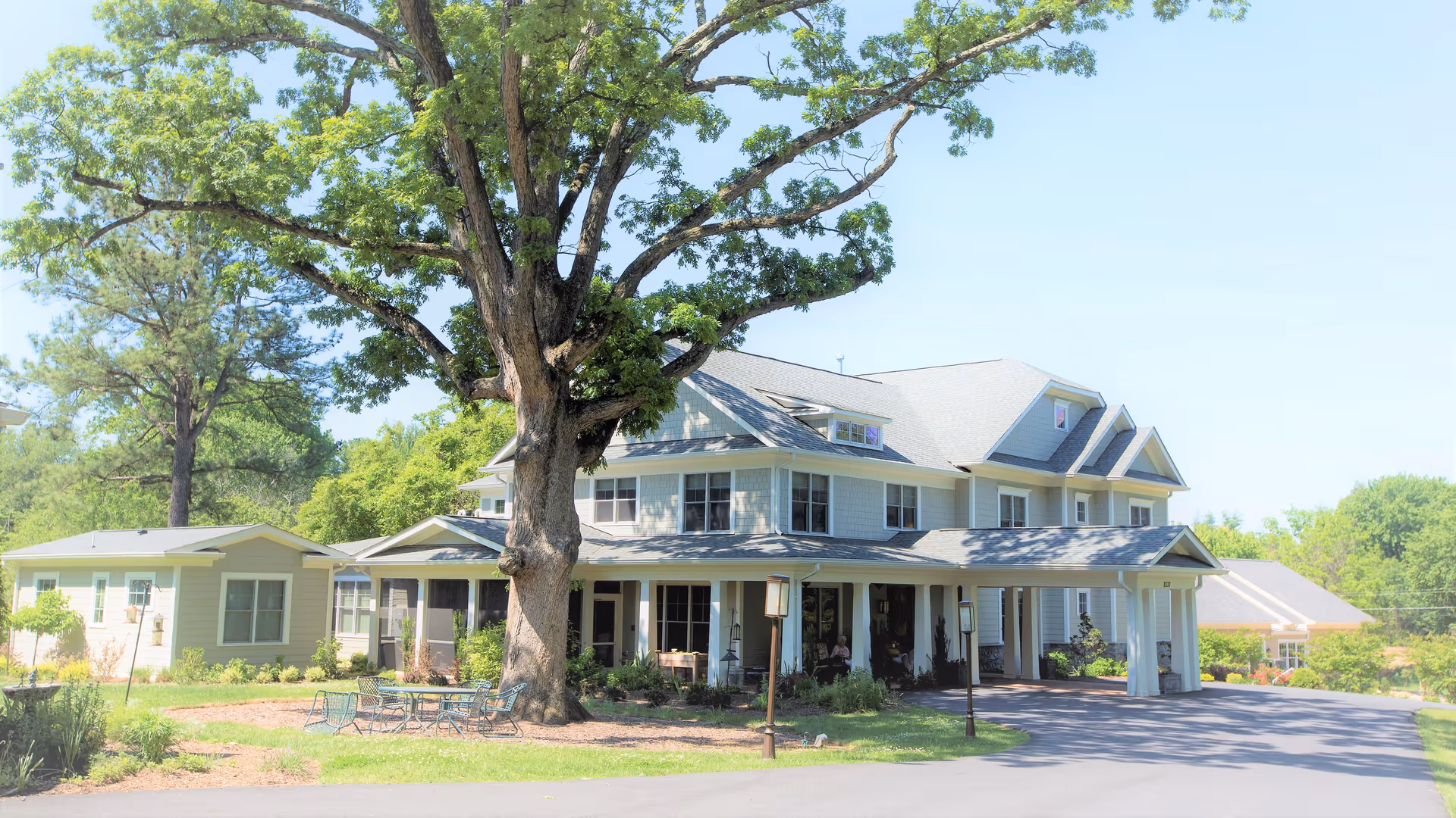 A large two-story residential building with a covered porch and multiple windows, surrounded by greenery and trees. There is a large tree in the foreground and outdoor seating with tables and chairs on the lawn. The sky is clear and blue.