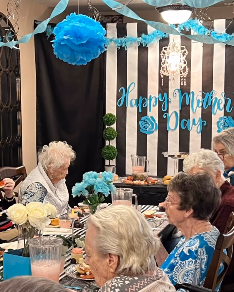 Elderly women seated around a decorated table celebrating Mother's Day with blue and white decorations and a 'Happy Mother's Day' backdrop.