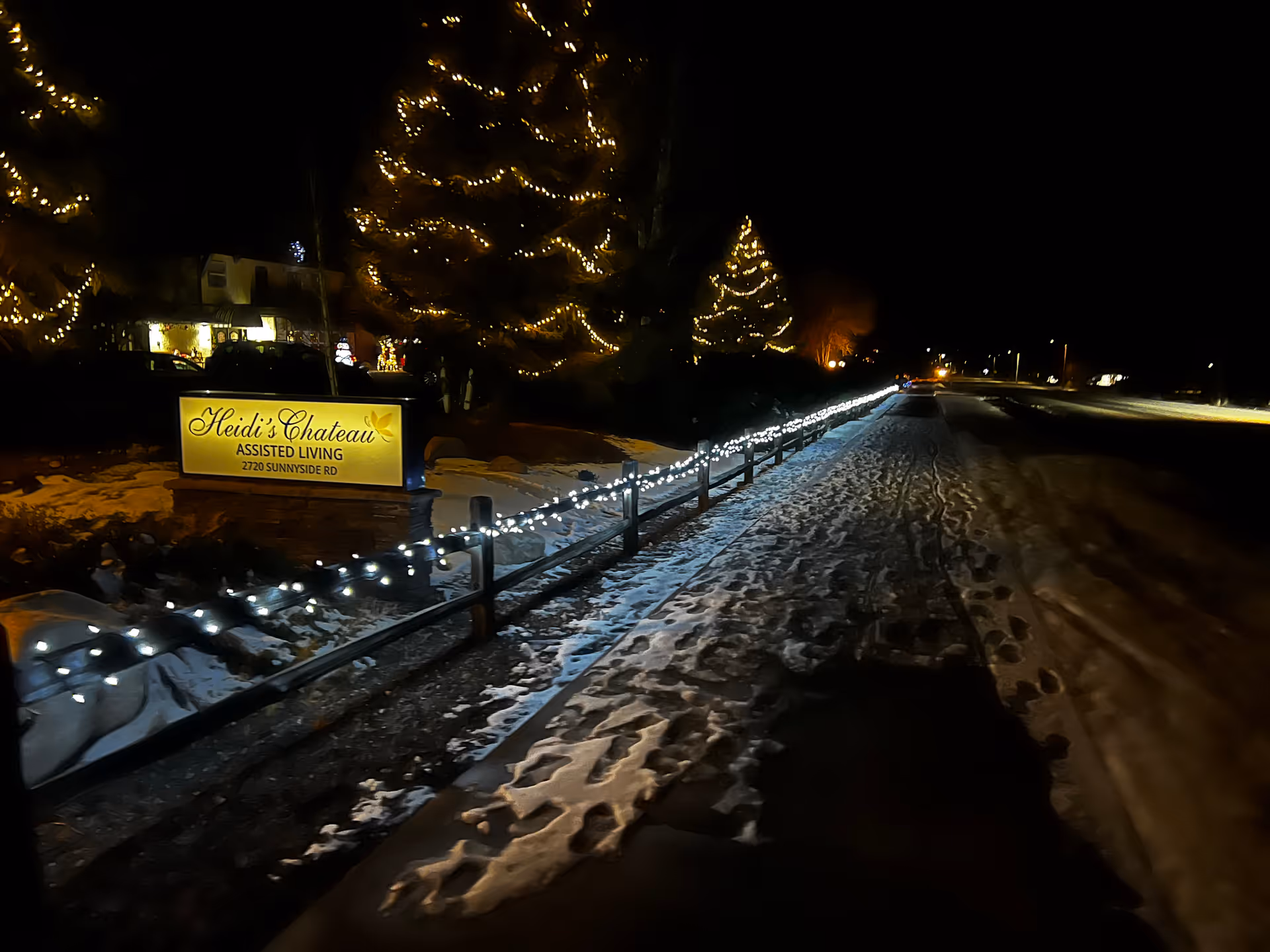 Night view of Heidi's Chateau assisted living sign with string lights on a fence, snow-covered walkway, and decorated trees.