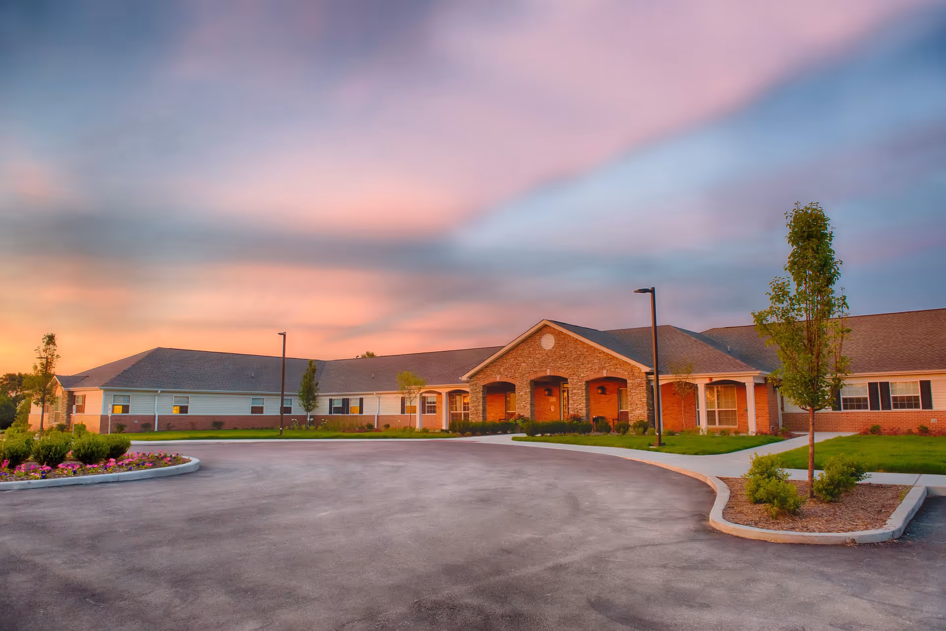 Exterior view of a single-story senior living facility building with a brick and siding facade, surrounded by landscaped greenery and a circular driveway under a colorful sunset sky.