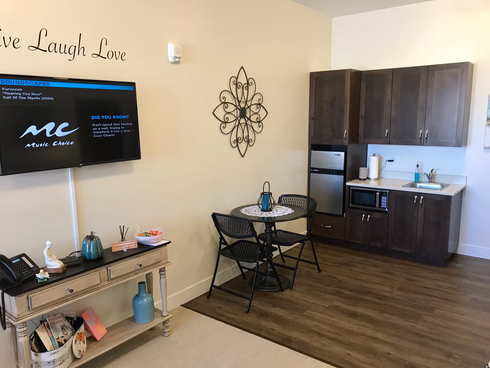 A cozy assisted living common area featuring a small kitchenette with dark wood cabinets, a mini refrigerator, microwave, and sink. There is a small round table with two black chairs on a wood floor. A wall-mounted TV displays a music channel, and a decorative metal wall art piece hangs on the beige wall. A console table holds decorative items, a phone, and books. The wall has the words 'Live Laugh Love' written on it.