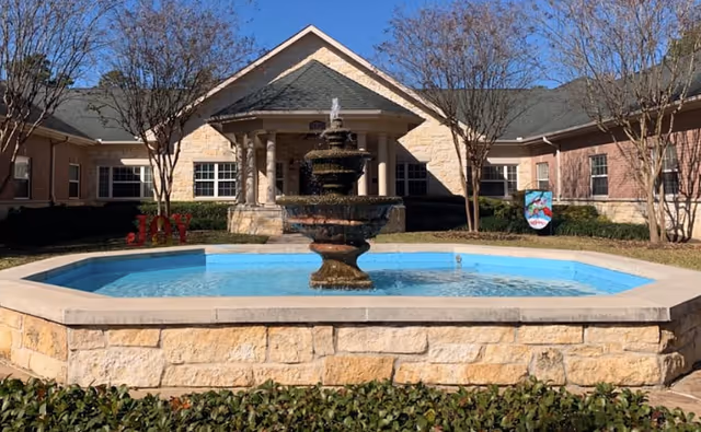 A multi-tiered stone fountain in a landscaped courtyard before the front entrance of a single-story brick building.