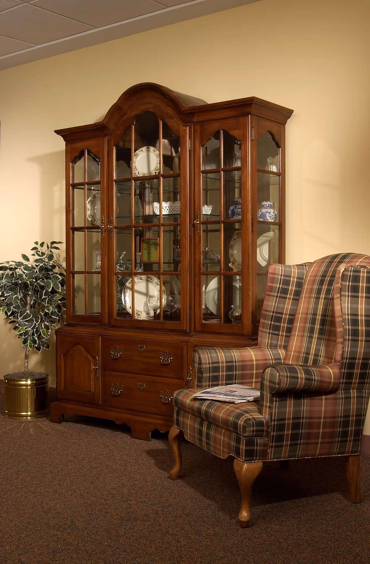 A cozy interior corner featuring a wooden china cabinet with glass doors displaying decorative plates and glassware, next to a plaid upholstered wingback chair with a newspaper resting on the seat. A potted plant in a brass container is placed beside the cabinet against a beige wall.
