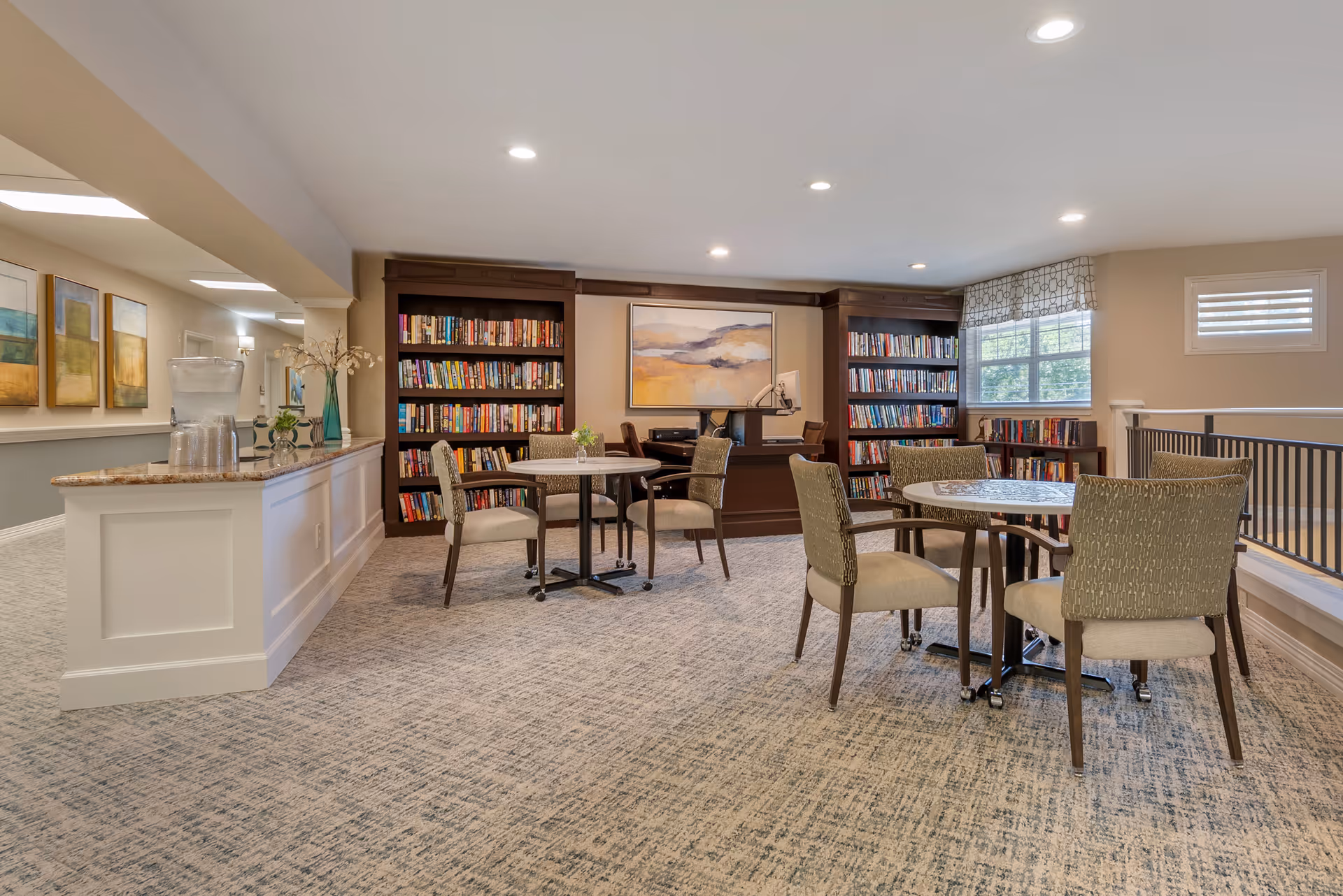 A bright and spacious reading or common area in a senior living facility with carpeted floors, several round tables with cushioned chairs, bookshelves filled with books, a large painting on the wall, and a window with a patterned valance letting in natural light. A counter with a water dispenser and decorative items is visible on the left side.