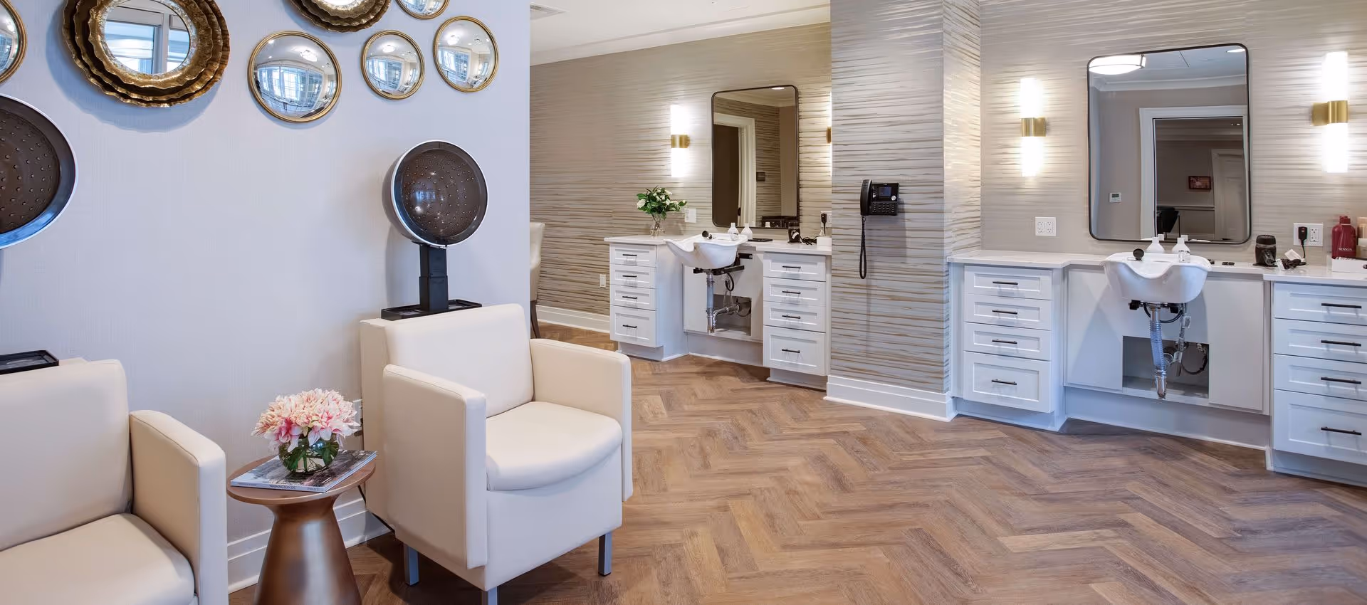 Interior of a salon area in a senior living facility with two beige armchairs, a small round table with a flower vase, two hair drying stations, and two white sink stations with mirrors and wall lights.