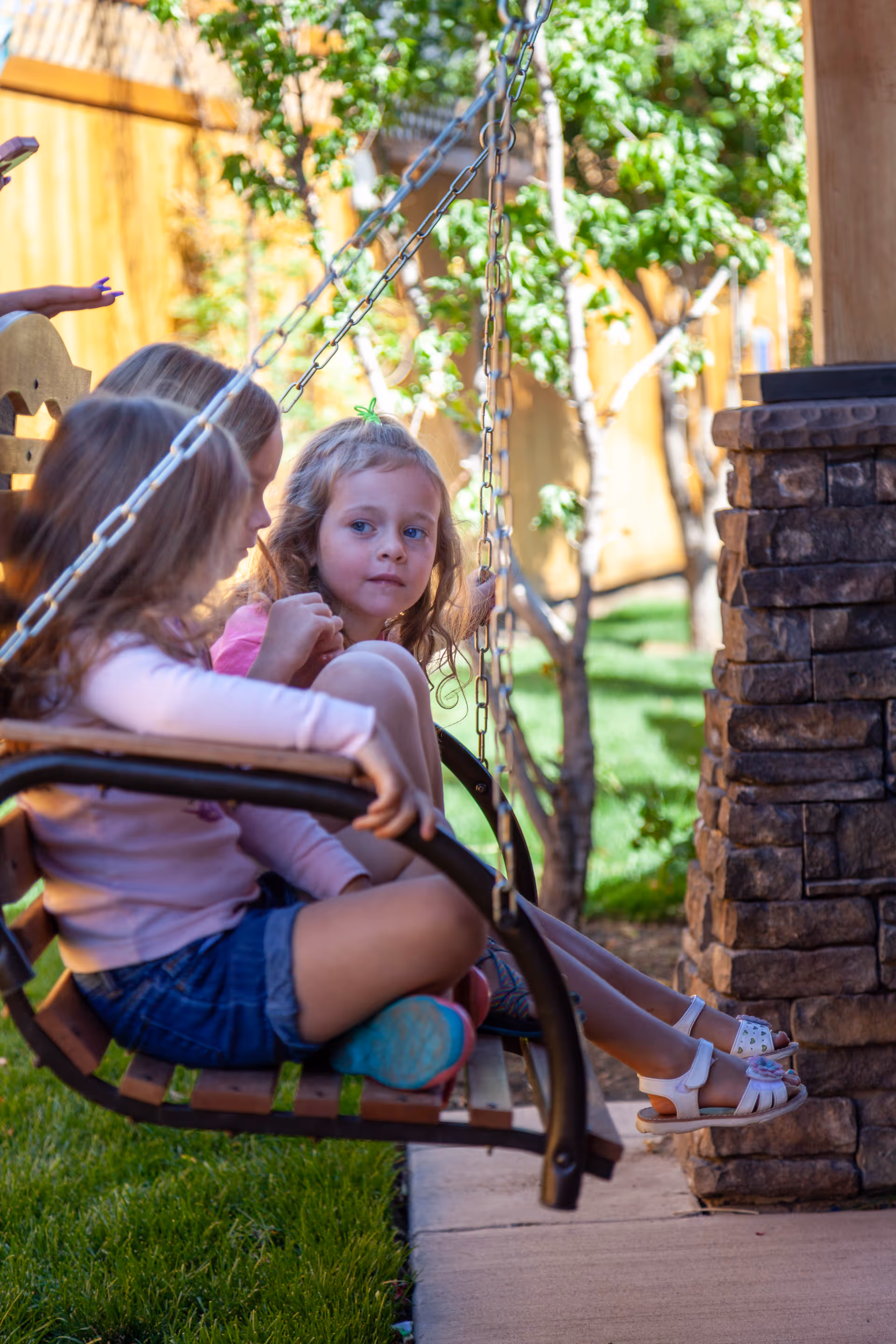 Two young girls sitting on a wooden swing with metal chains in a garden area, with green grass, trees, and a stone pillar visible nearby.