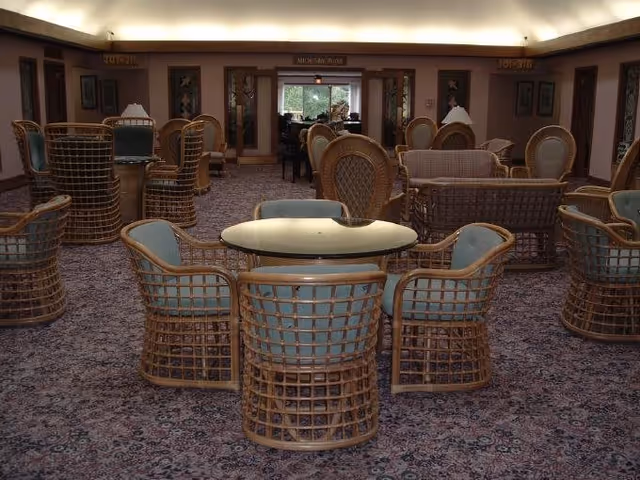 A spacious common area with multiple wicker chairs and tables arranged on a patterned carpet. The room has soft lighting and framed artwork on the walls. There is a sign above a doorway in the background that reads 'Welcome Home.'