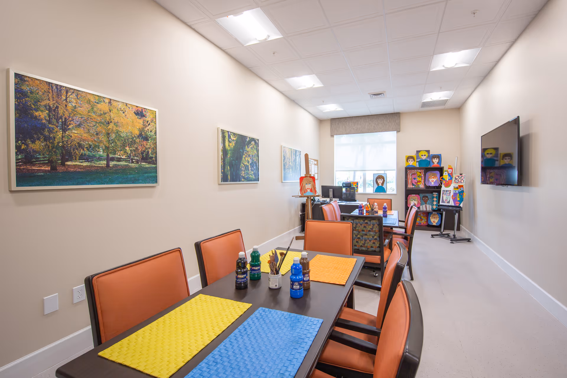 A bright room with beige walls and ceiling lights, featuring two tables with orange chairs around them. The tables have colorful placemats and bottles of paint or similar materials. The far end of the room has an easel, shelves with colorful paintings of faces, and a large window with a shade. There is a large flat-screen TV mounted on the right wall and framed landscape pictures on the left wall.
