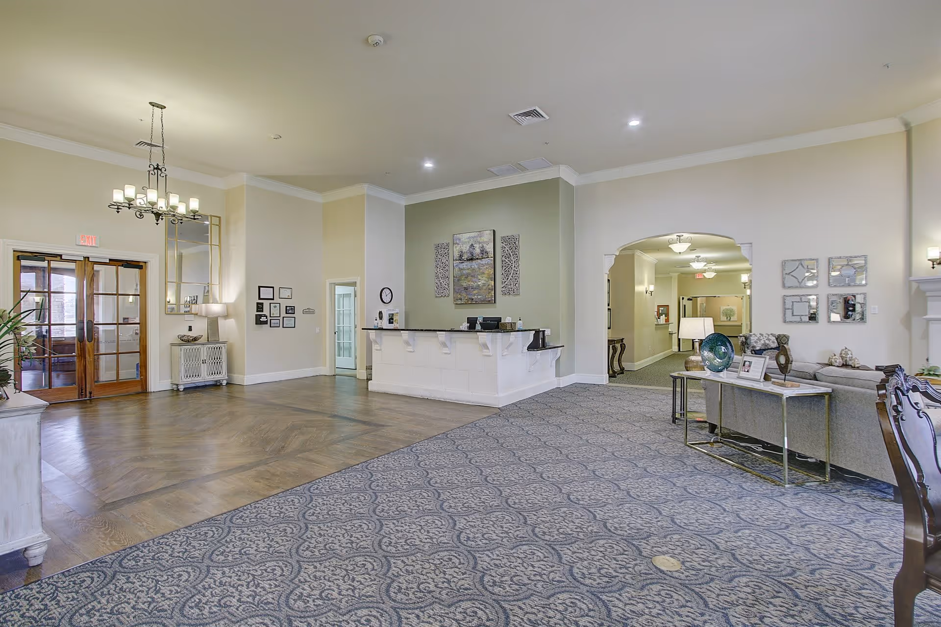 A spacious senior living facility lobby with a reception desk against a light green accent wall. The floor is partially carpeted with a patterned design and partially wooden. There is a chandelier hanging from the ceiling, a seating area with a sofa and table on the right, and double glass doors on the left. The walls are painted in neutral tones with decorative artwork and mirrors.